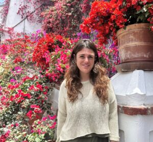 Board member Jessi, a woman with long brown hair wearing an off-white sweater standing outdoors with autumn trees in the background