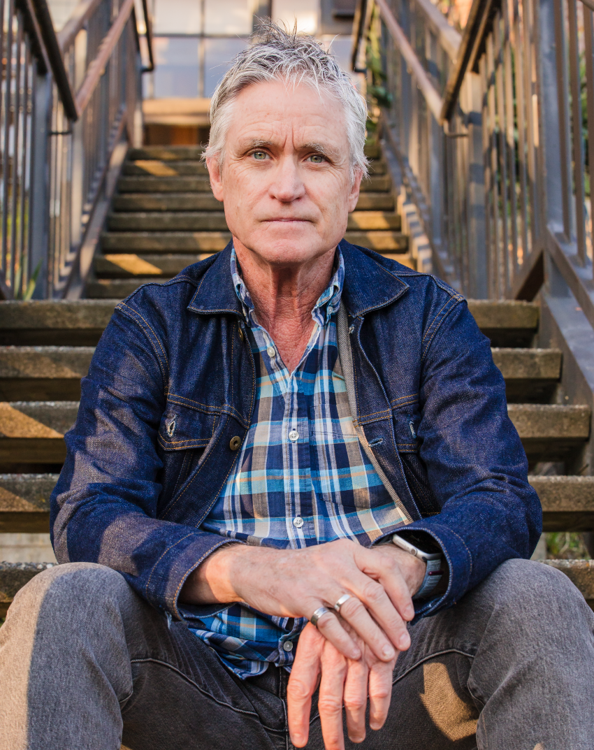 Tom is a an older white male with short grey hair wearing a denim jacket and plaid shirt sitting at the bottom of a wooden staircase