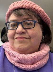 White woman with a full, round face with mid-length brown hair wearing a pink beret, pink knit scarf and purple glasses.